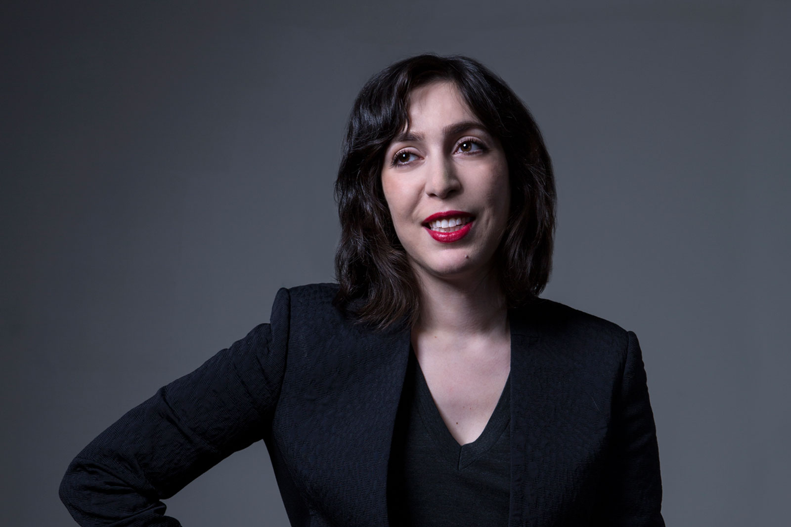 A woman with shoulder-length dark brown hair wears a black blazer over a dark v-neck top. She has red lipstick and her right hand rests on her hip. The background is a solid dark gray.