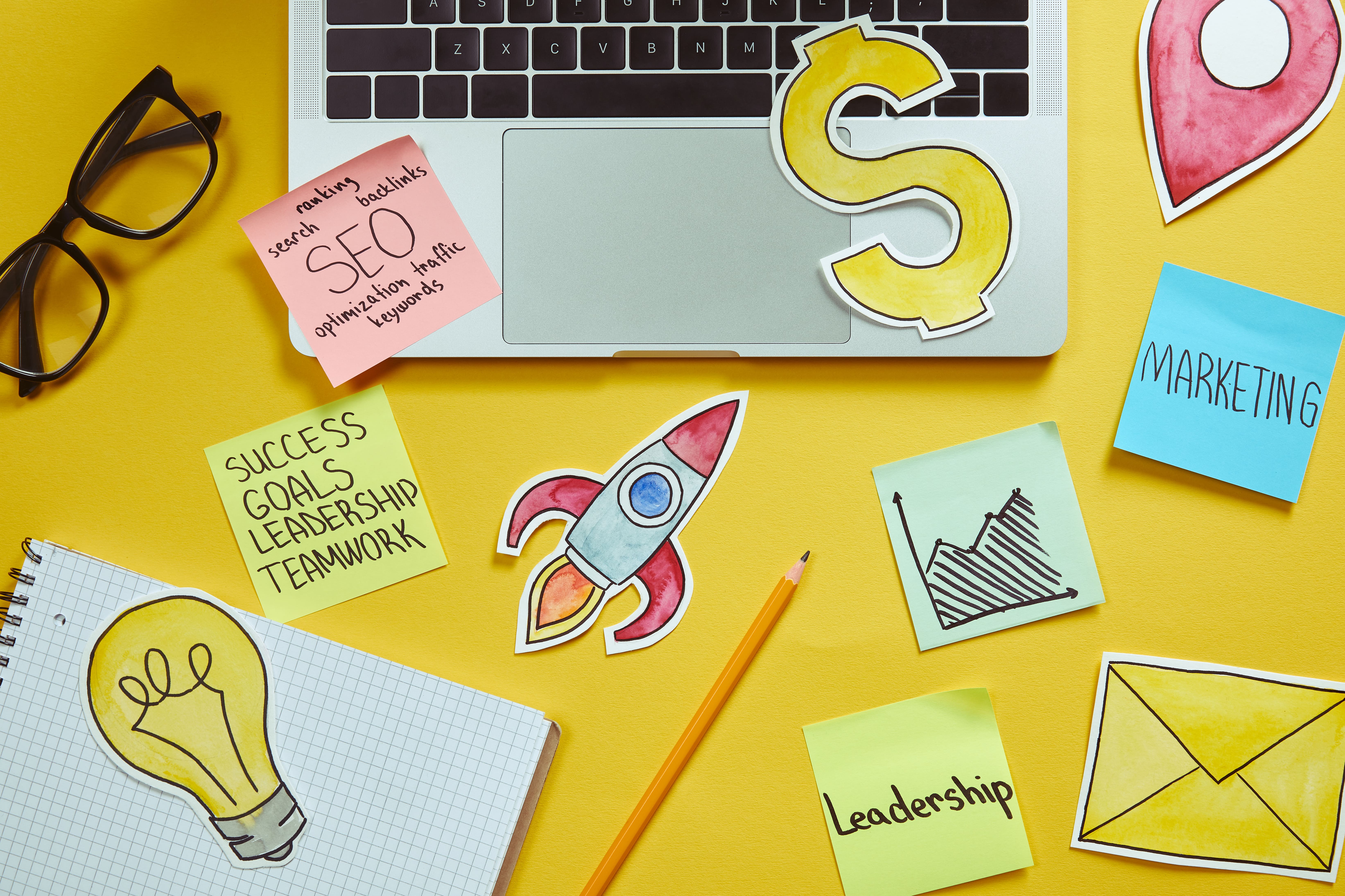 Top view of laptop with SEO, marketing, leadership, and business strategy concept elements including sticky notes, rocket, light bulb, and dollar sign on yellow background