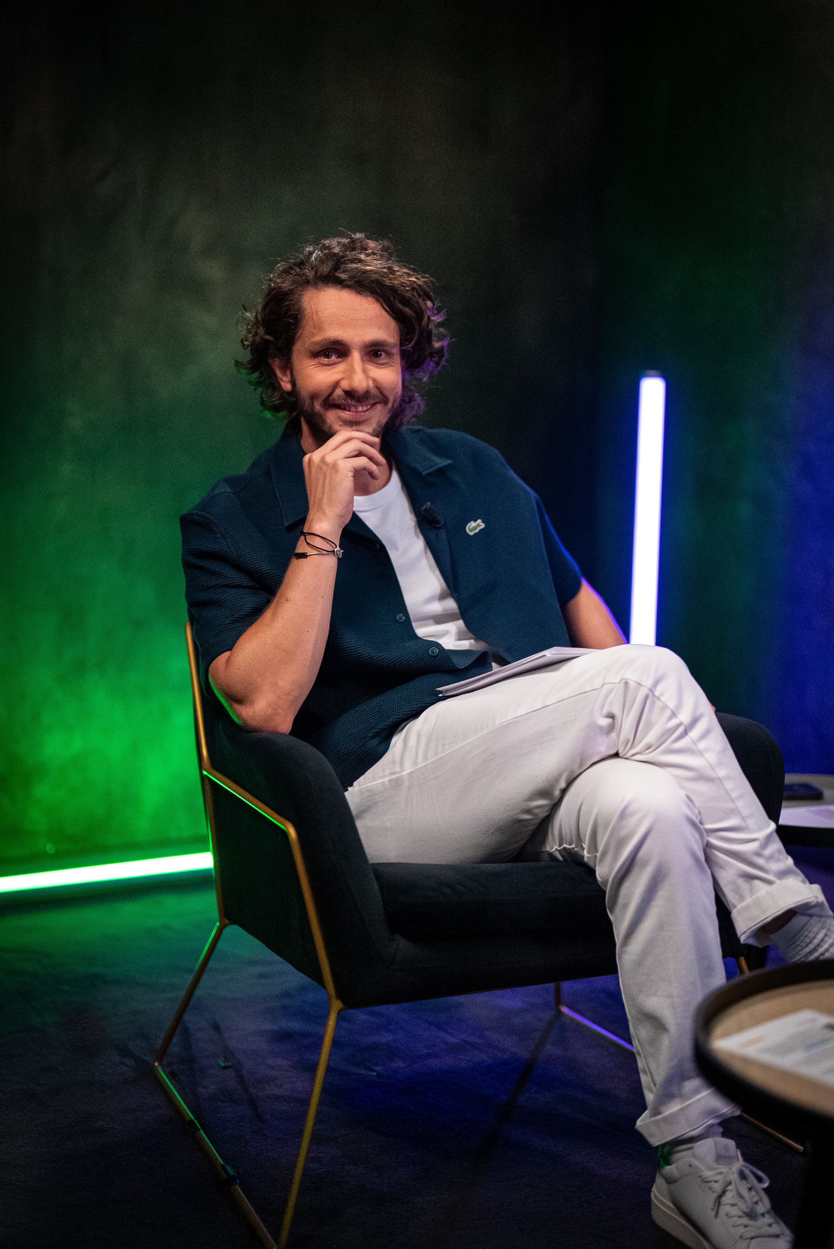 Photographie professionnelle de Guillaume Pley (fondateur Legend) posant sur un fauteuil moderne dans son studio. Il sourit, la main sous le menton, vêtu d'une chemise bleu marine décontractée et d'un pantalon blanc. L'image est caractérisée par un éclairage de studio créatif (lumière néon verte et bleue en arrière-plan), typique des productions de médias numériques. Cette photo de haute qualité, optimisée par retouche et post-production, est un exemple de service Photo & Vidéo fourni par Nux-Studio pour la création de contenu de marque et le branding de célébrités.