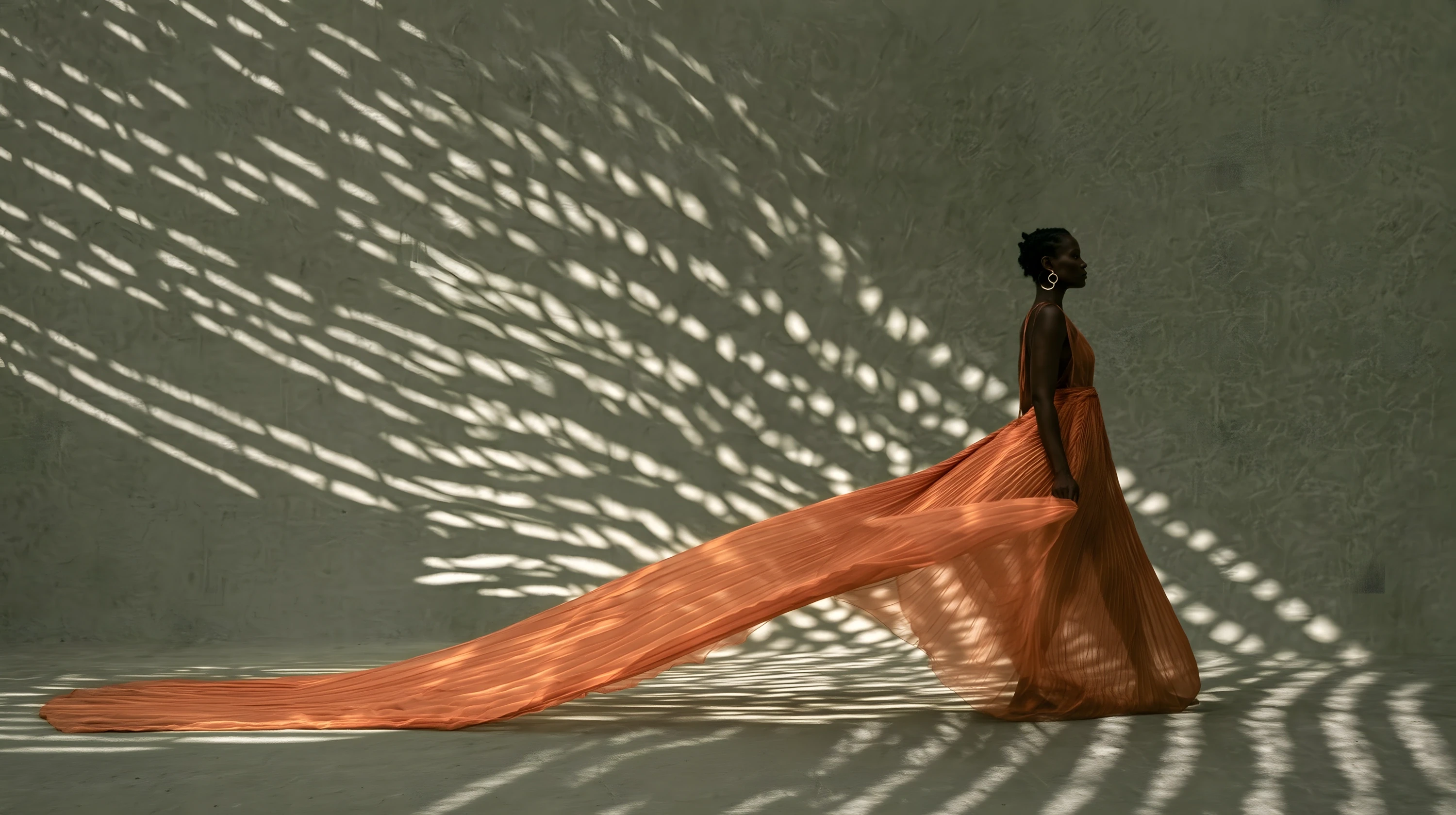 	Woman in a flowing terracotta gown standing against a sage wall patterned with dappled light