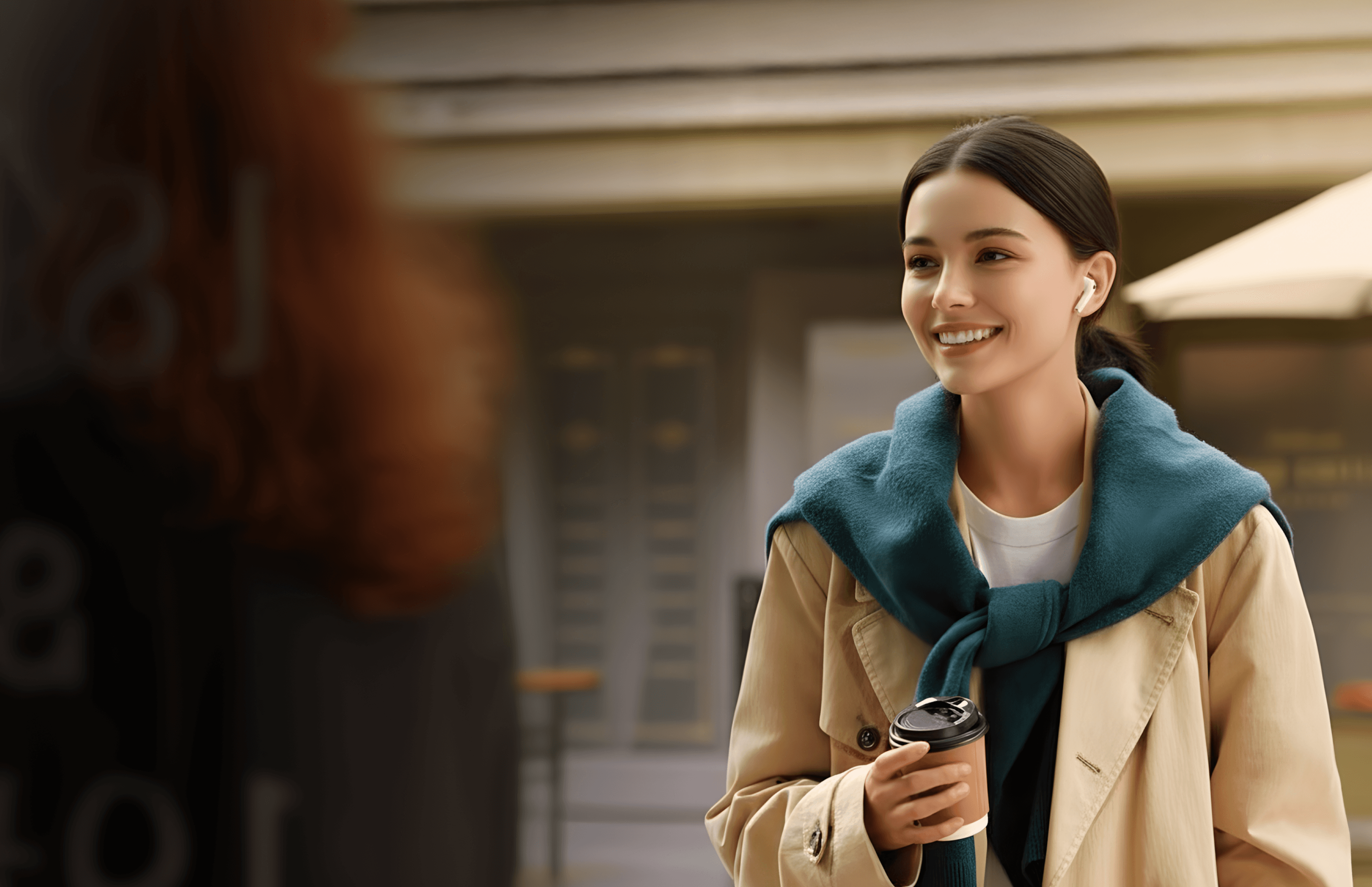 Smiling woman with earbuds, wearing a blue scarf and trench coat, holding coffee, walking on a city street.