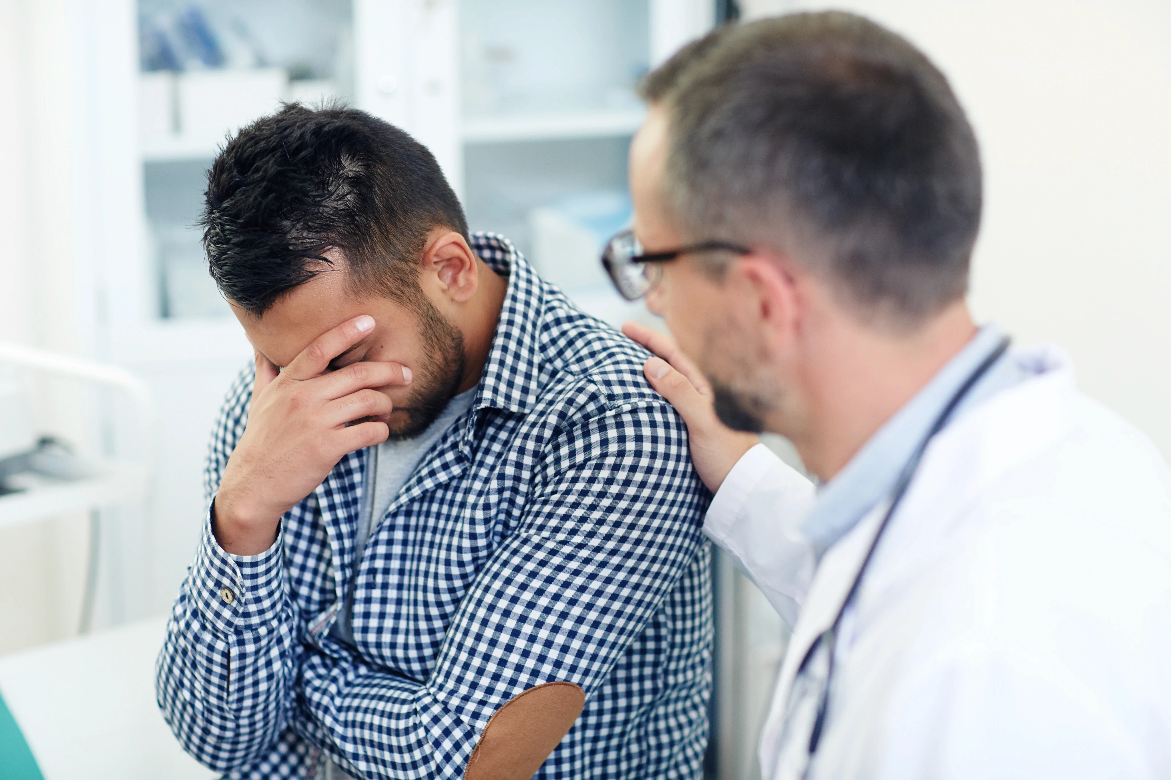 A doctor consoles a distressed man in a blue checkered shirt by placing a hand on his shoulder, conveying empathy and support in a clinical setting.