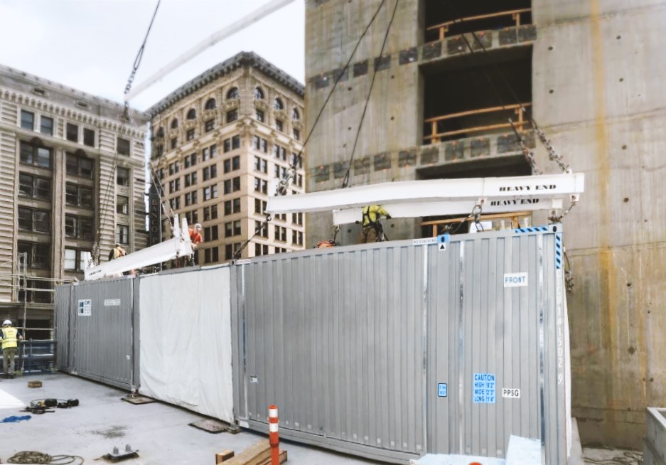 A crane lowers a guestroom module onto a concrete podium during construction.