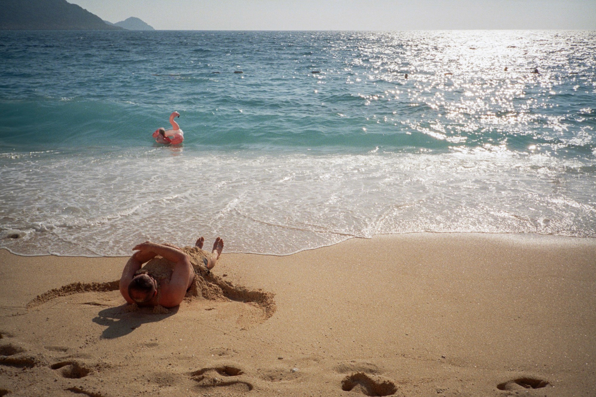 A man is buried in sand on a sunlit beach, with gentle waves lapping the shore and another person wading in the turquoise ocean.