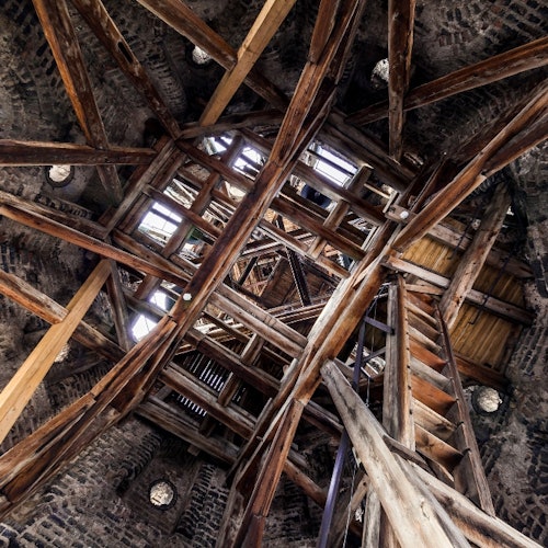 View of wooden beams and ladders forming a complex structure within a stone tower. Light enters through openings above.