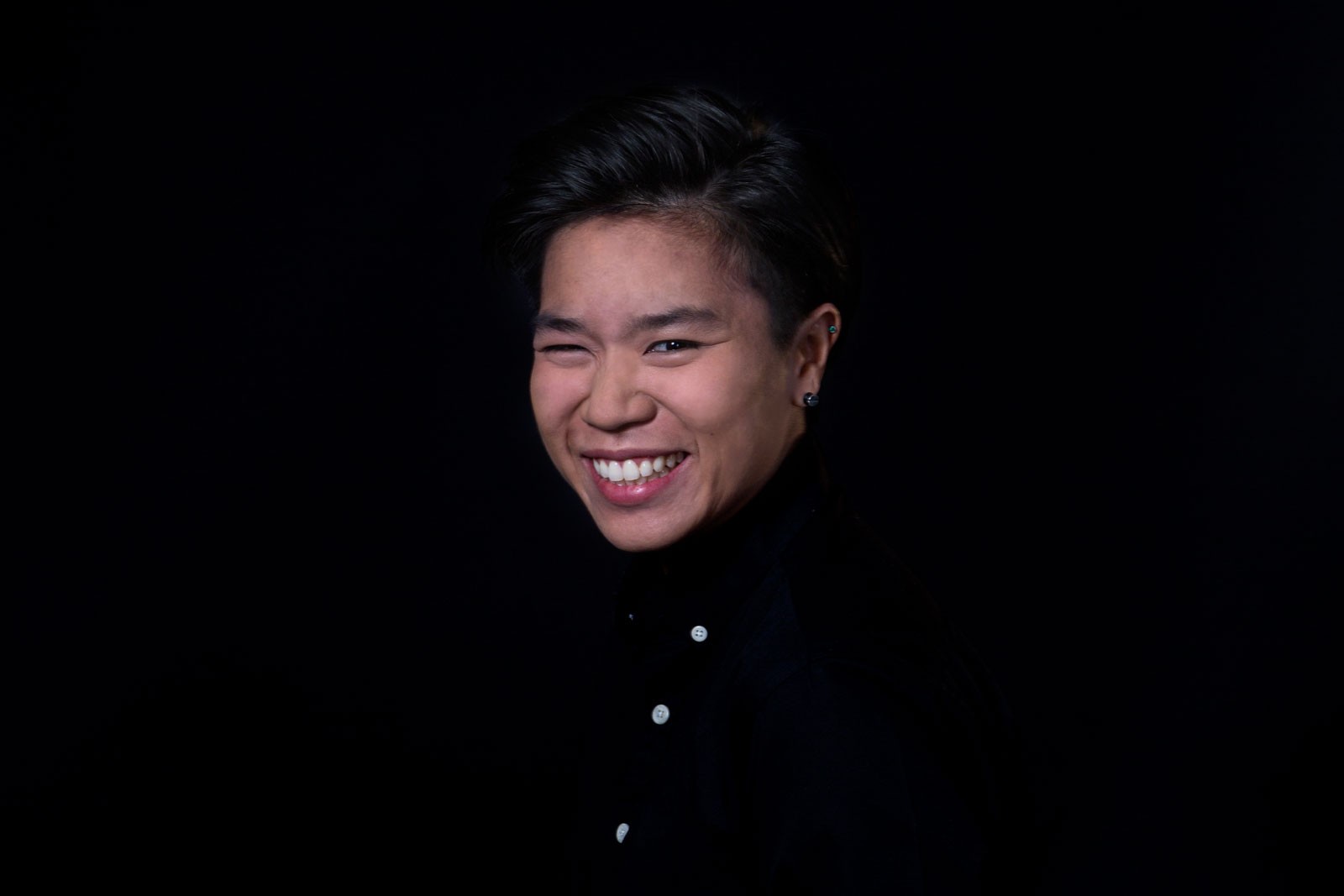 A person with dark hair and dark eyes is smiling broadly against a solid black background. They are wearing a dark-colored shirt with three visible buttons near the collar. The individual’s head is turned slightly to the left, and their mouth is open in a wide smile, revealing their teeth. The lighting is focused on the person's face, leaving the rest of the image dark and indistinct.