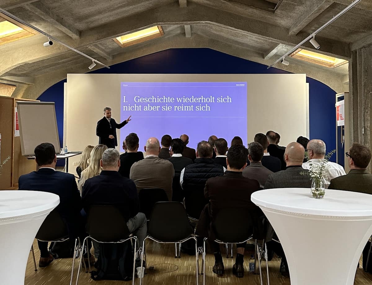 Vortragsszene in einem modernen Raum mit Leinwand, Stehtisch und Stuhlreihen; zwei Personen präsentieren ein digitales oder technologisches Thema.