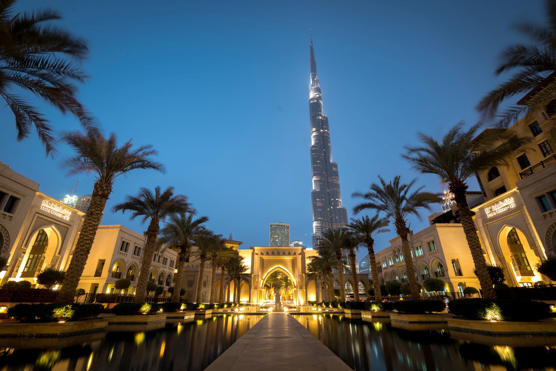 The Burj Khalifa tower rises above a luxury resort courtyard with palm trees and lit fountains at twilight.