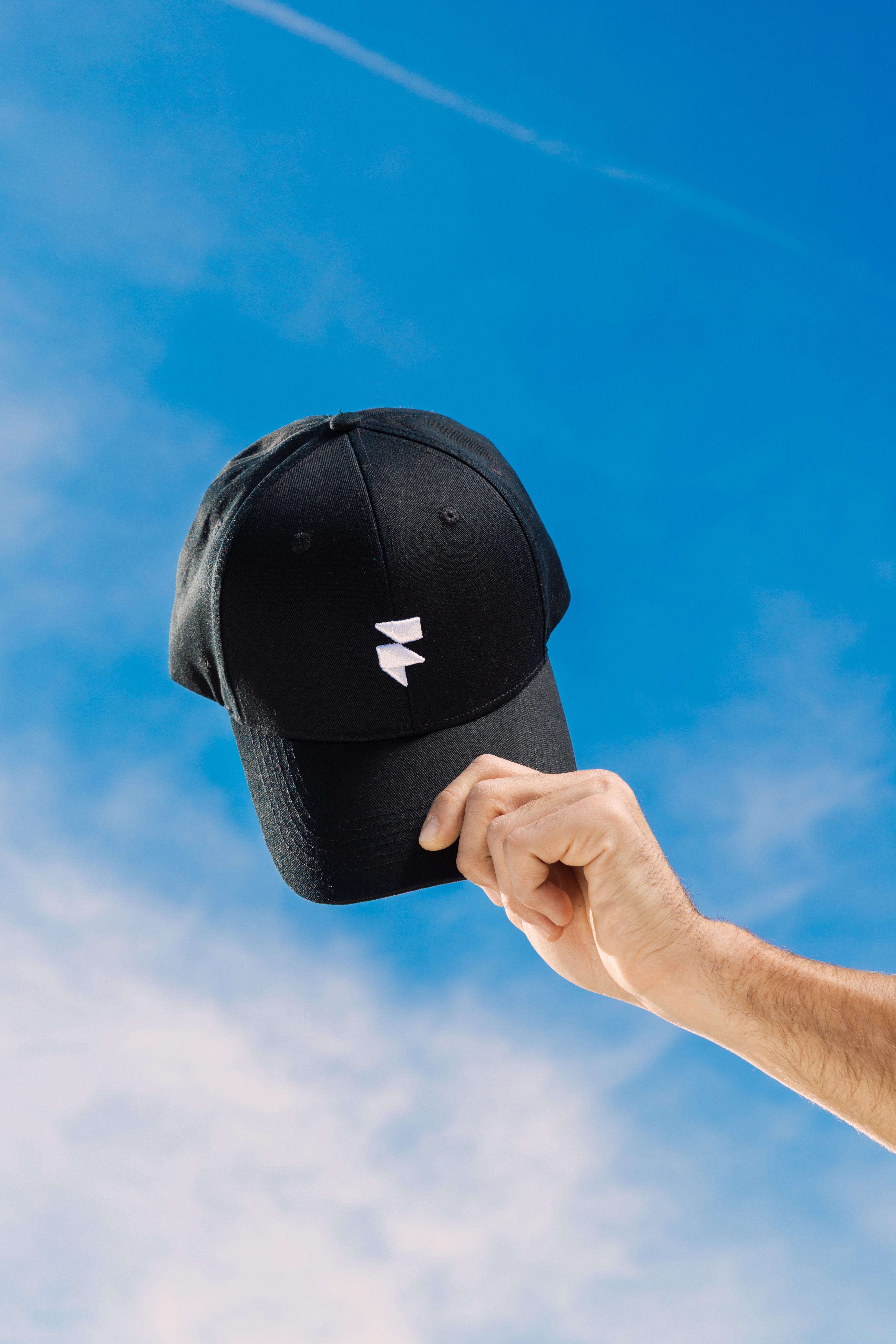 A hand holds up a black baseball cap against a vibrant blue sky with wispy clouds, showcasing the cap's white emblem prominently.