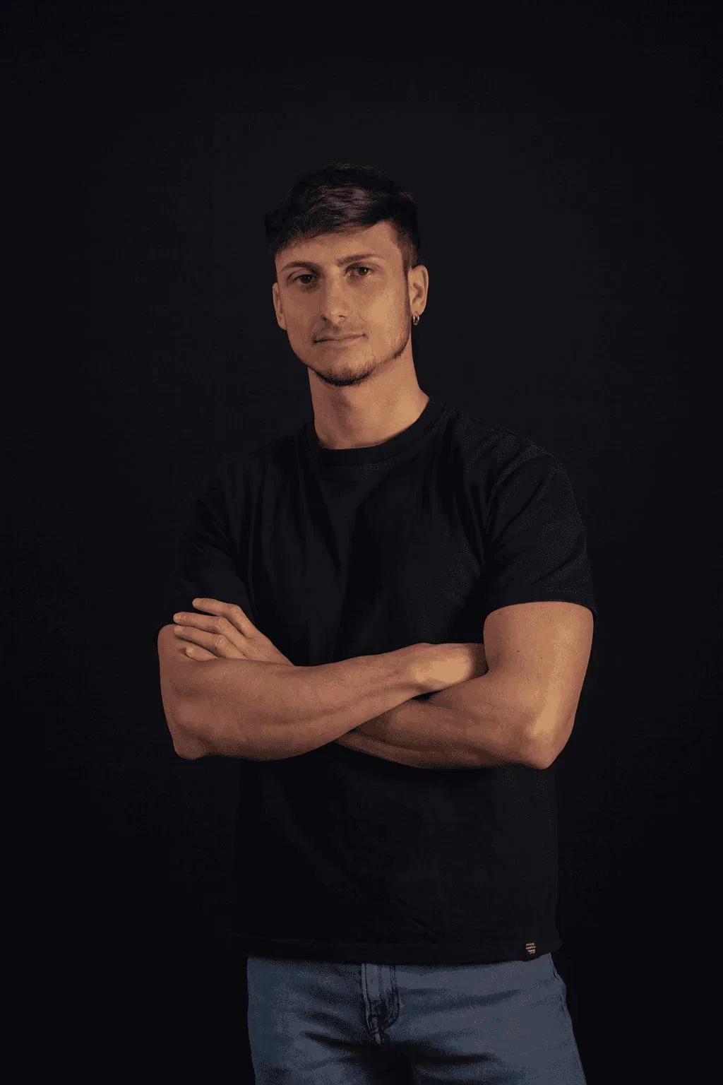 Young man with dark hair and stubble, black tee, arms crossed.