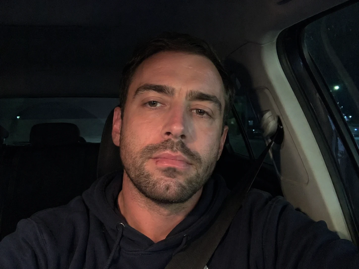 Portrait of a man sitting in a car at night wearing a dark hoodie with low ambient lighting