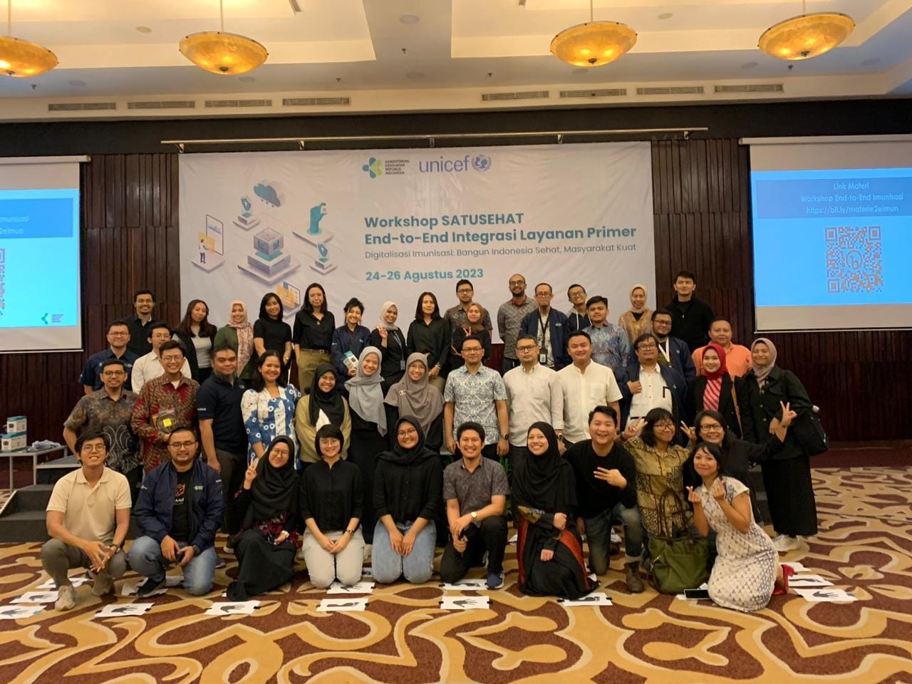 Forty participants grouped on a carpeted stage in a hotel ballroom beneath a banner reading ‘Workshop SATUSEHAT End-to-End Integrasi Layanan Primer, 24–26 Agustus 2023’.