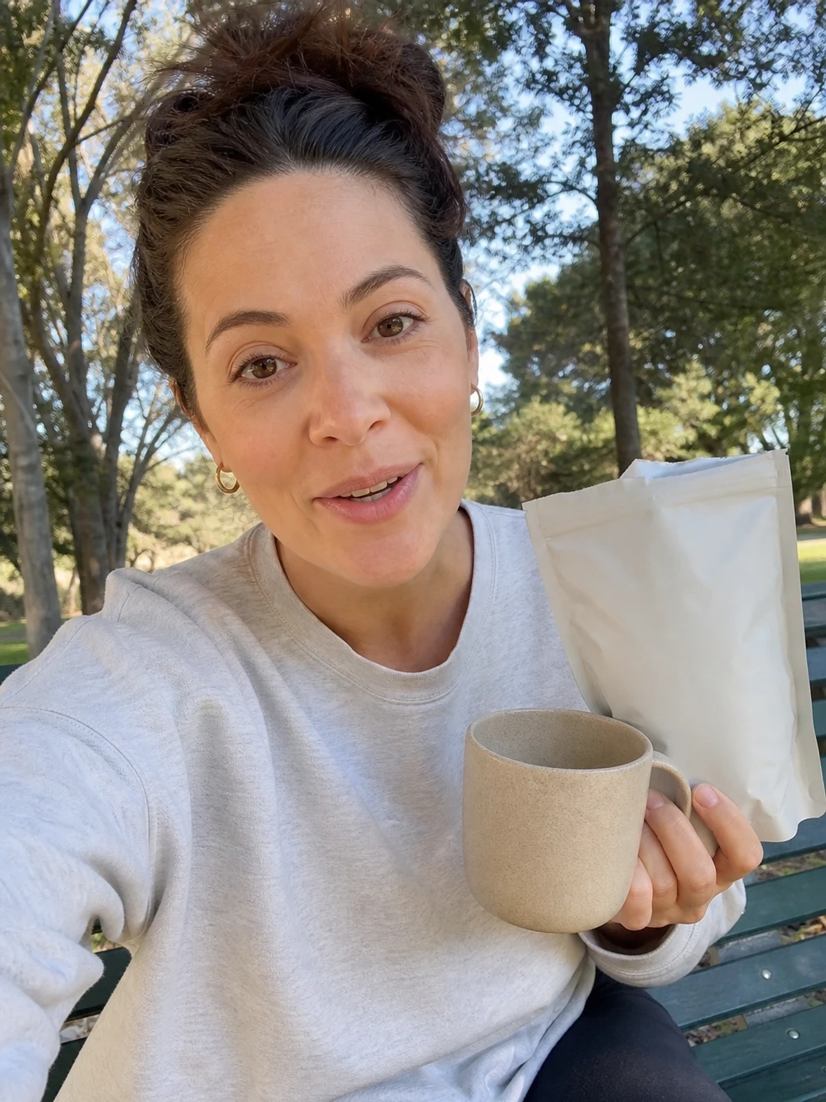 UGC-style photo: parent creator, on a park bench, showing a coffee pouch and mug while talking to camera, selfie angle.