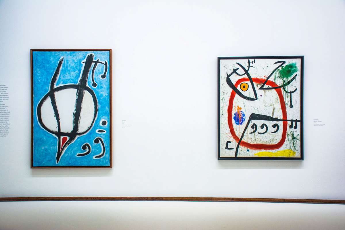 Two abstract paintings with bold lines and primary colors hang on a white gallery wall above a wooden baseboard.