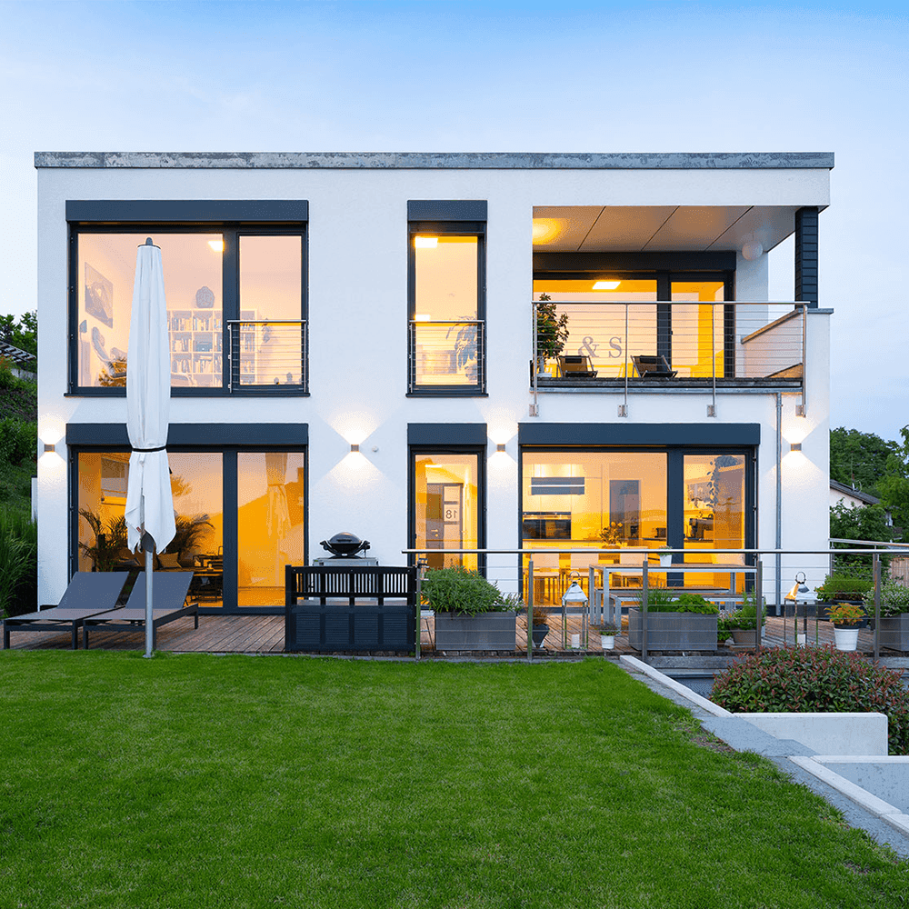 Moderne weiße Villa mit großen Fenstern, Balkon und Terrasse in der Abenddämmerung, beleuchtet von Außenleuchten.