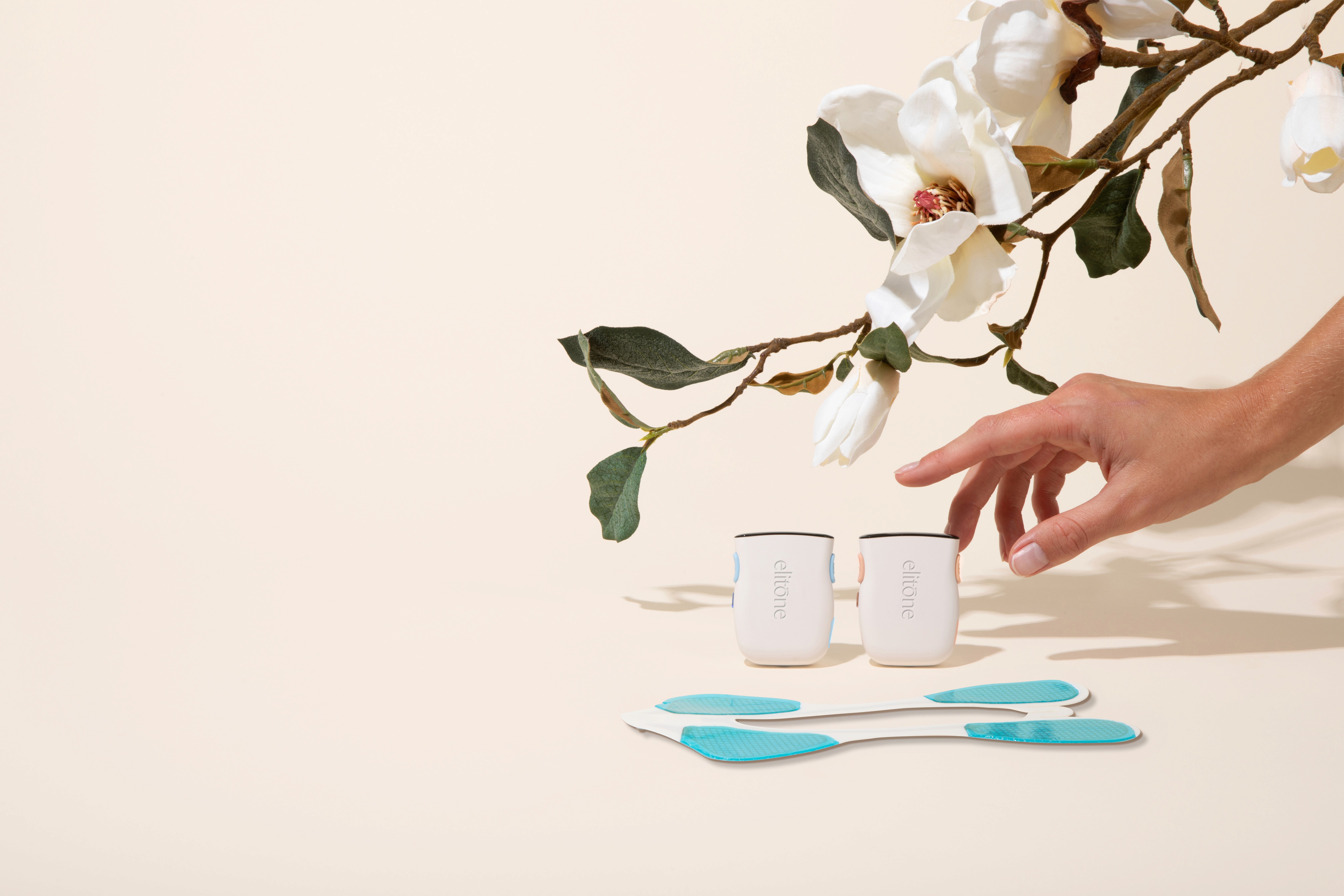 A hand reaches for two small white cups beneath a magnolia branch, with blue utensils in the foreground.