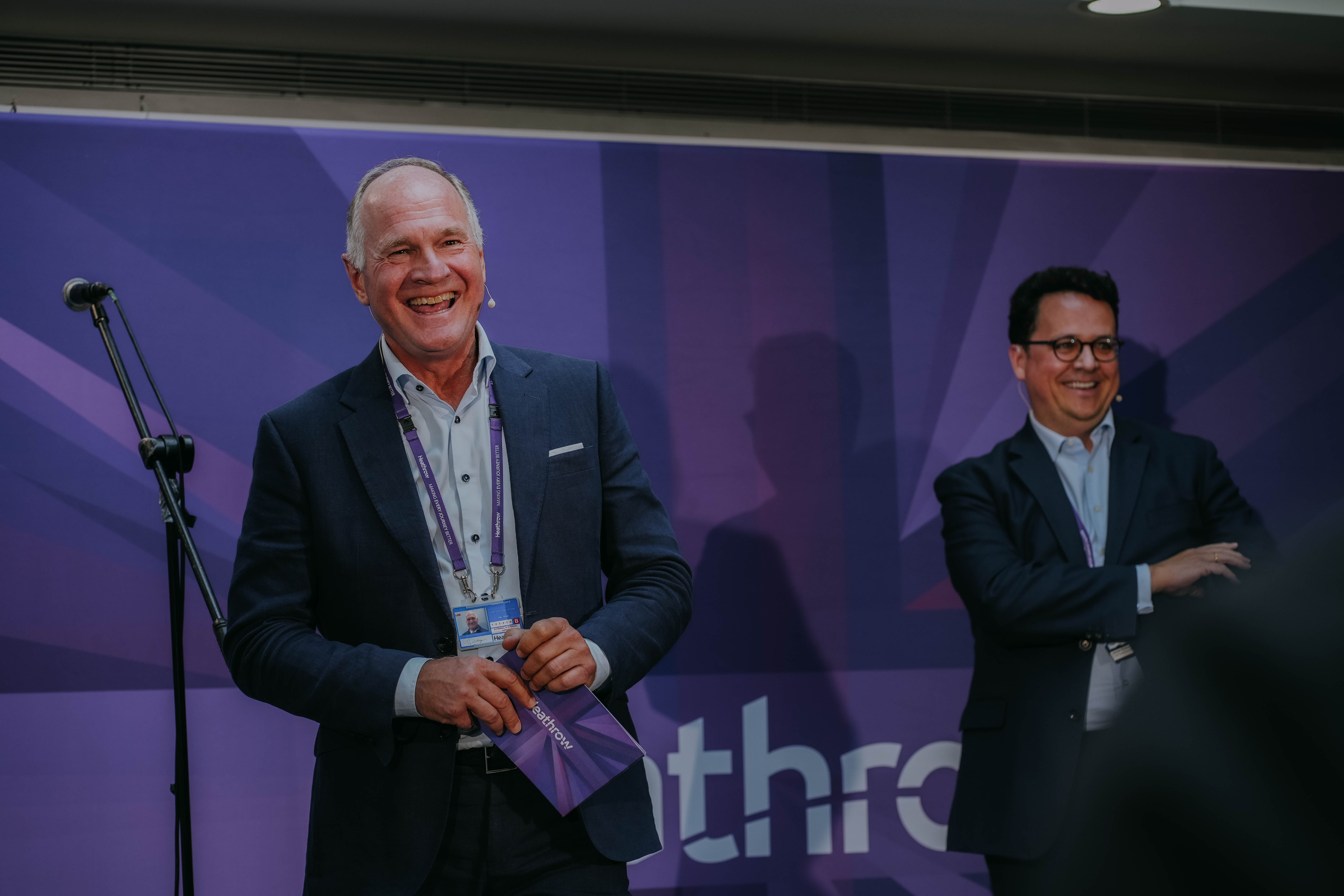 Heathrow CEO Thomas Woldbye (left) laughs heartily on stage while holding a pamphlet. To his right, Heathrow COO Javier Echave smiles with his arms crossed. They are in front of a large purple banner.