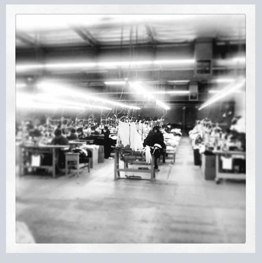A high-angle, intentionally blurred black-and-white photograph of a large commercial garment sewing floor, showing rows of industrial apparel manufacturing equipment and operations.