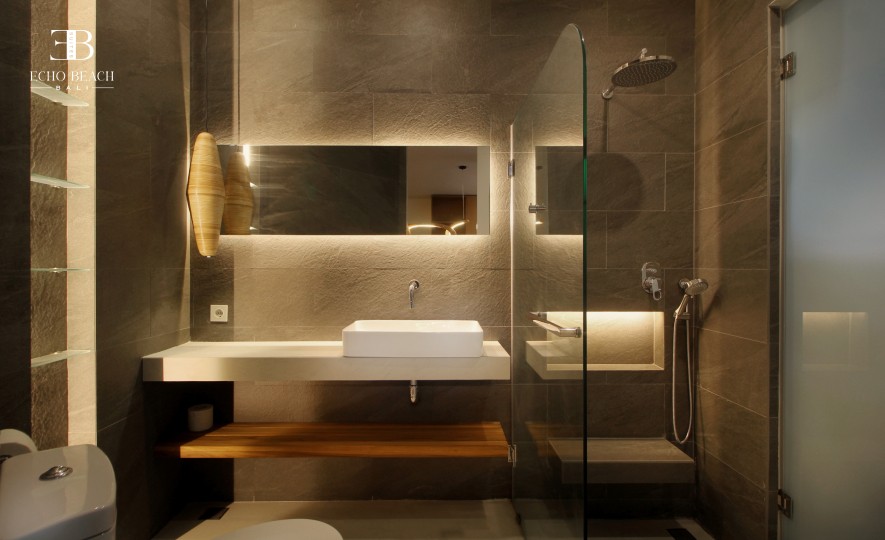 Full view of the luxurious bathroom with dark stone tiles, a glass-enclosed rain shower, floating vanity, and wooden shelf at the Echo Beach Suite, located near Echo Beach in Canggu, Bali.