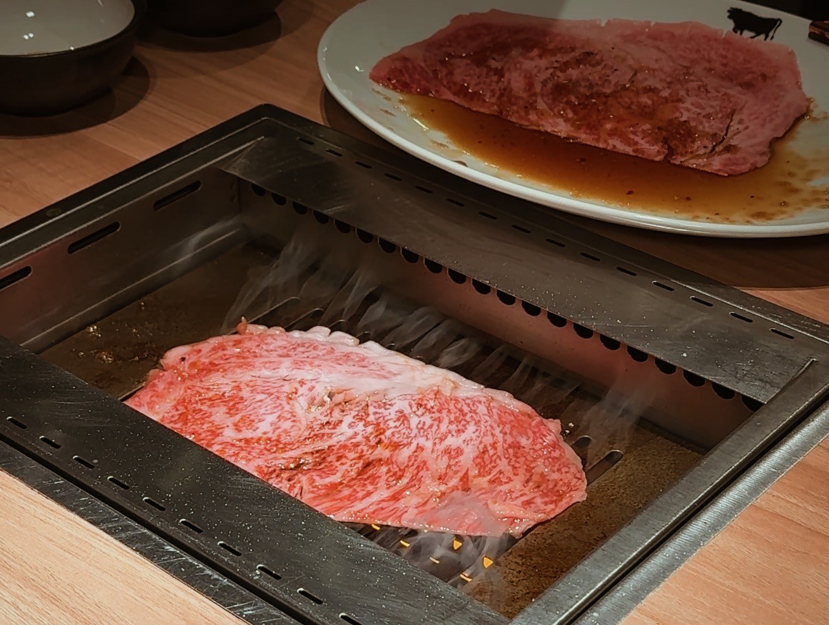 A piece of marbled beef sizzling on a tabletop grill, with another slice of raw beef on a plate in the background.