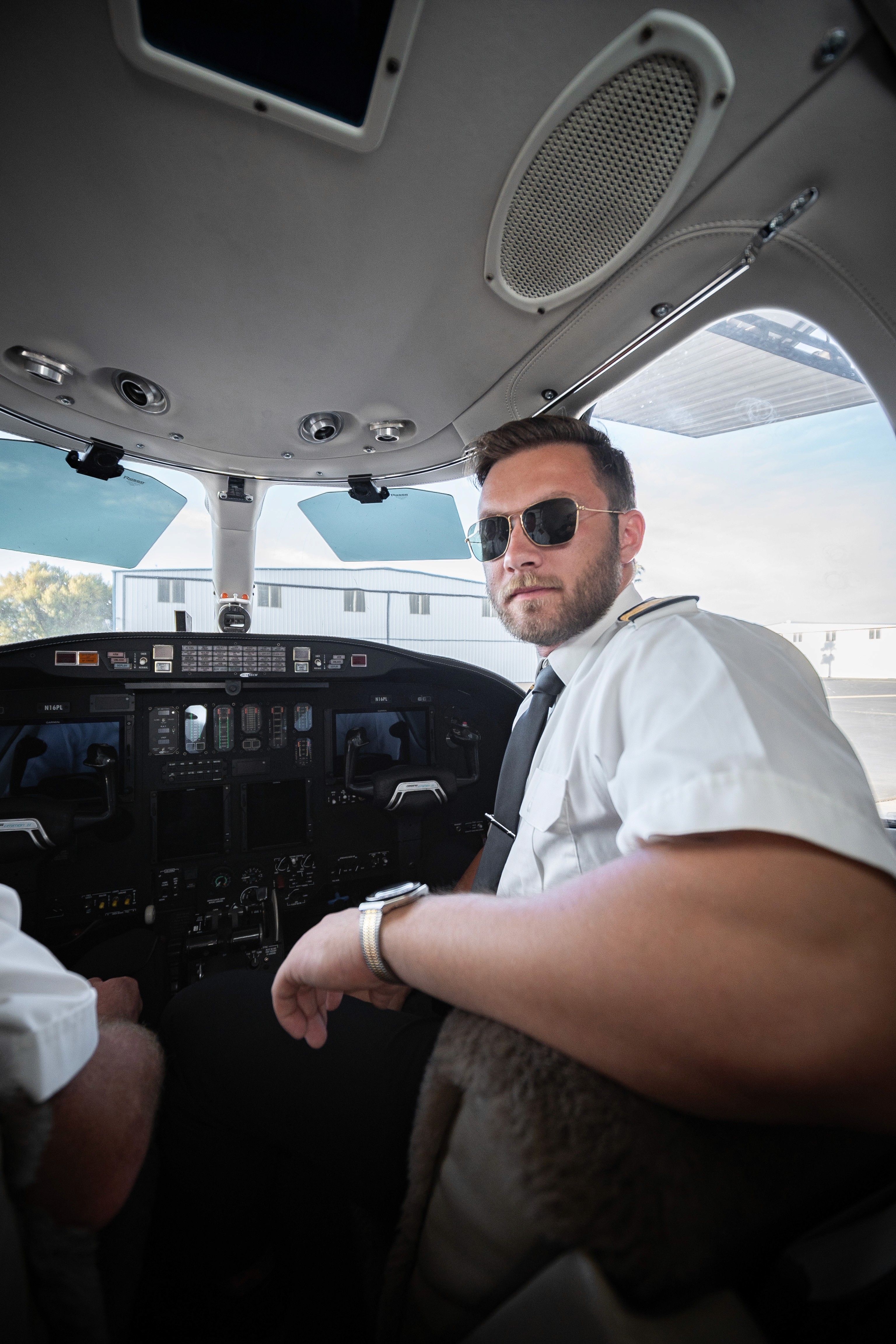 Smiling man wearing a beige overshirt and jeans, seated against a light wooden background.