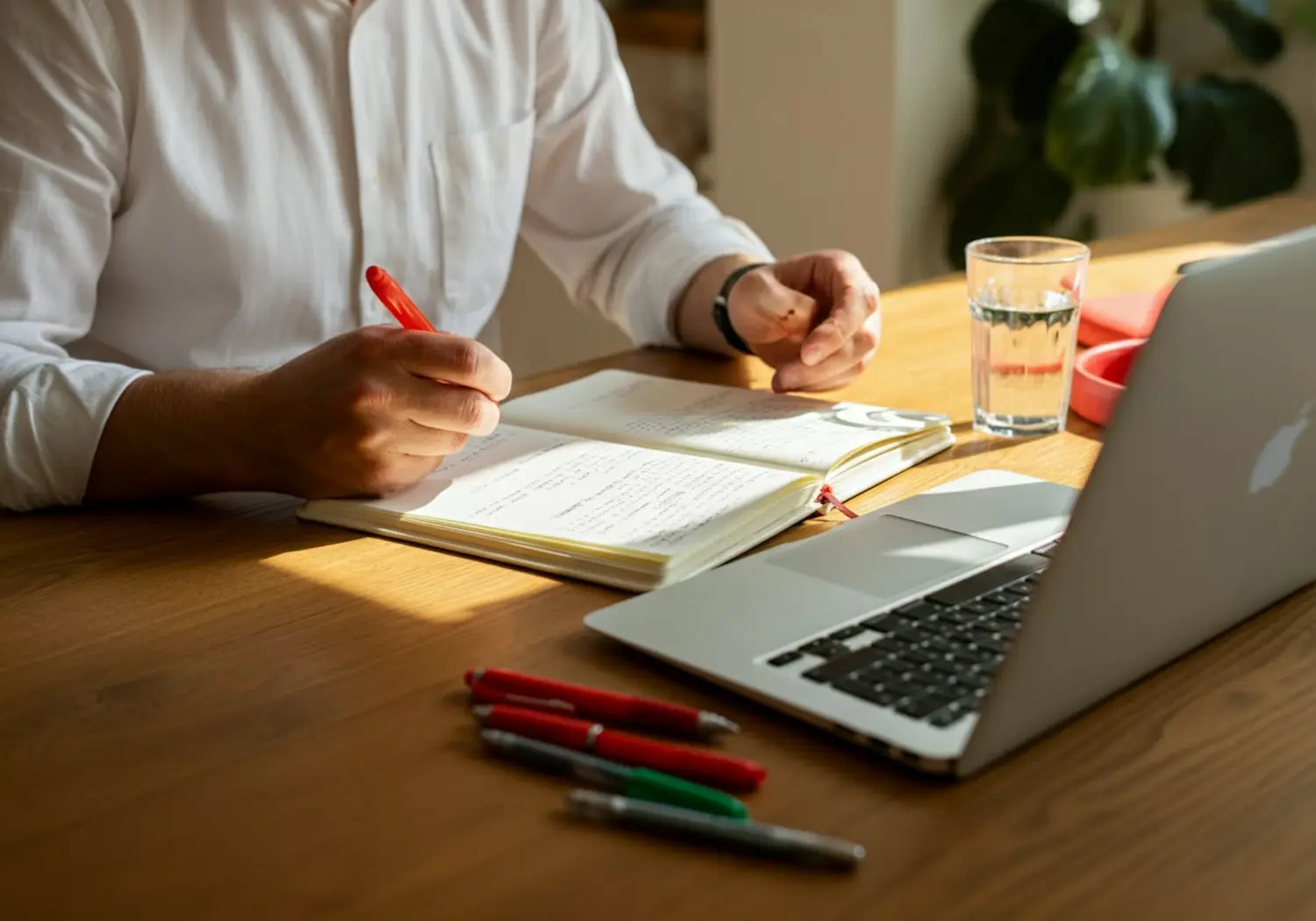 Person writing in a notebook beside a laptop on a wooden desk.