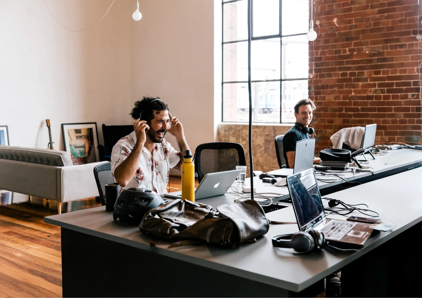 A modern office environment with two people working at a long desk, featuring laptops, headphones, and personal items, set against a backdrop of large windows and exposed brick walls.