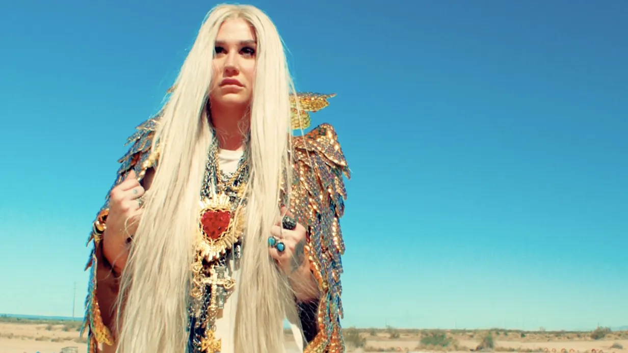 A person with long white hair stands in a desert landscape, wearing traditional attire and jewelry against a clear blue sky.