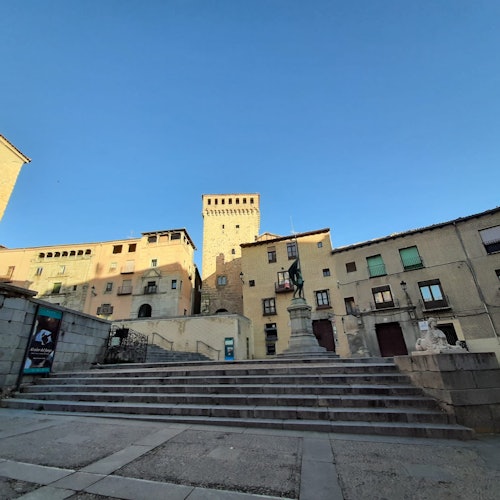 Stone steps leading to a plaza with a central statue, historic buildings, and a clear blue sky in the background.