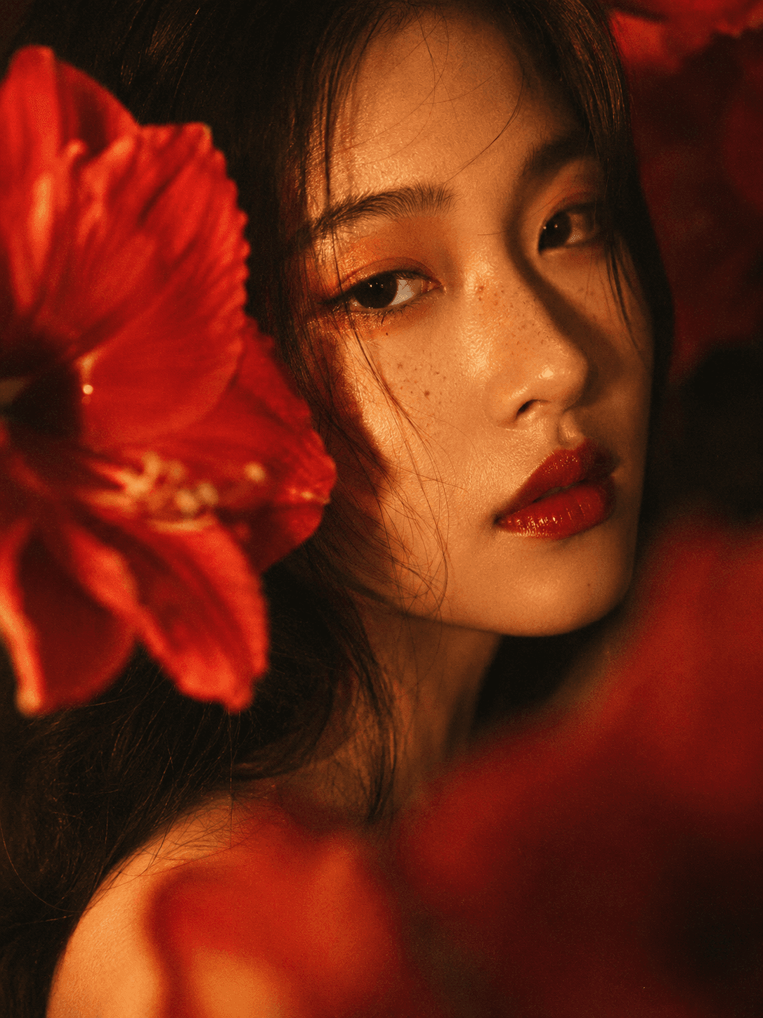 Close-up of a woman with bold red makeup and freckles, surrounded by red flowers. Warm lighting casts shadows, creating a dramatic, vibrant tone.