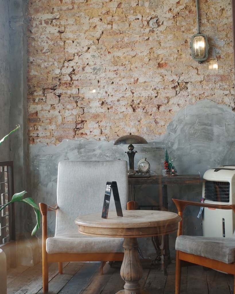 Cozy café interior featuring a vintage wooden table, two soft chairs, a small decorative tree, and a rustic brick wall.