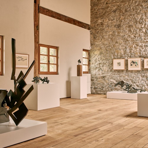 艺术画廊内部设有现代雕塑矗立于白色底座上，石墙上挂有装裱的艺术作品，地板为木制材质。