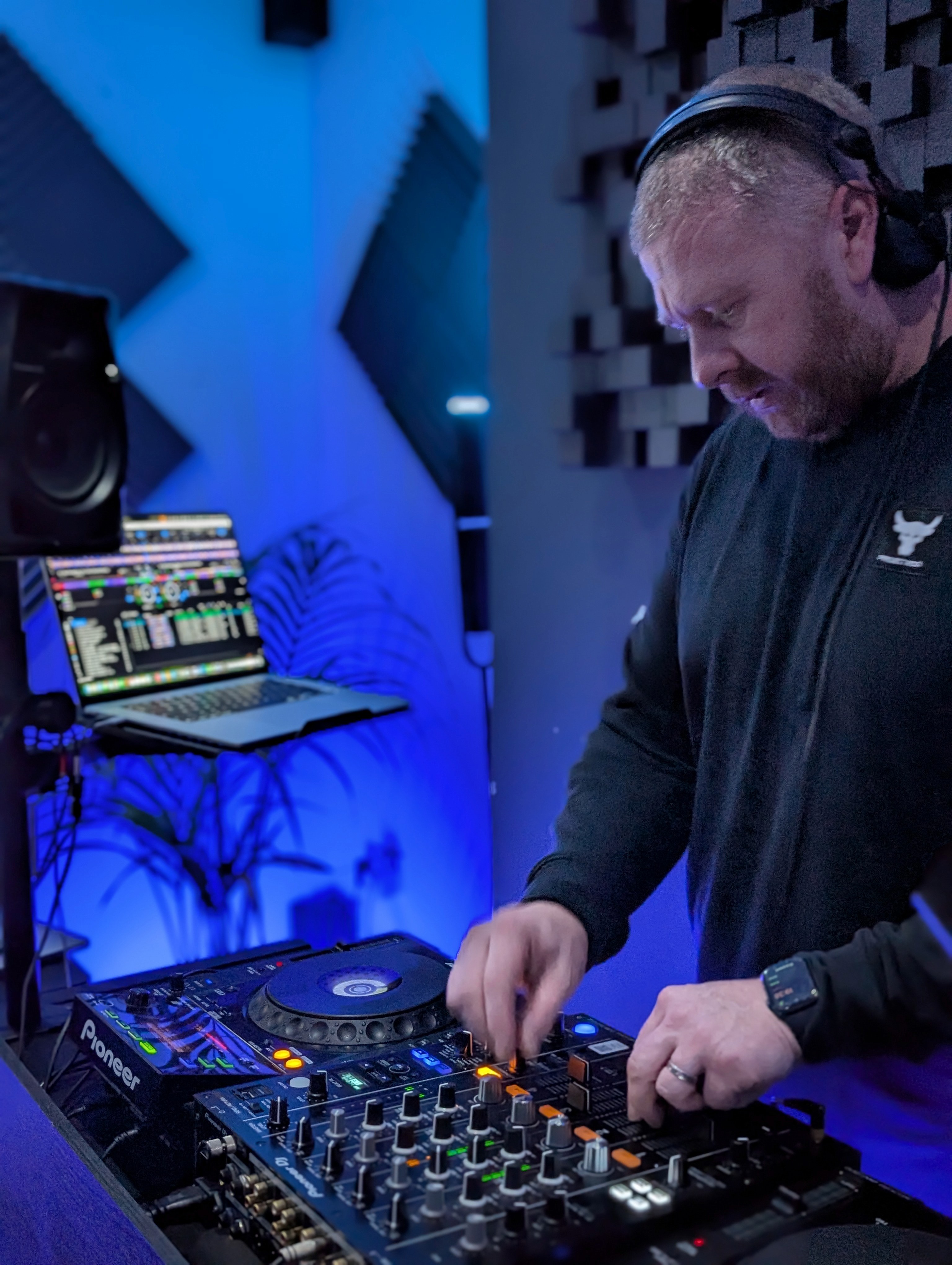 A DJ standing at a mixing board, wearing headphones and a white t-shirt, working with Pioneer DJ equipment.
