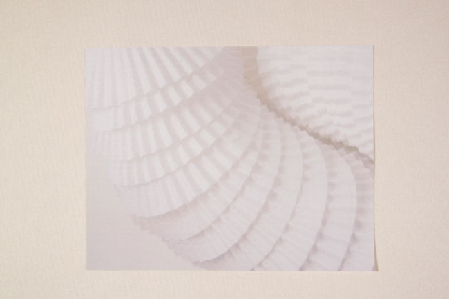 The image shows a piece of white paper with a series of overlapping, pleated or folded sections arranged in concentric circles. The paper is positioned against a light beige or cream-colored background. The design is monochromatic and features smooth, curved lines that create a sense of depth and movement. The paper’s surface has a slightly textured appearance due to the pleats. The image is framed by a thin dark border.