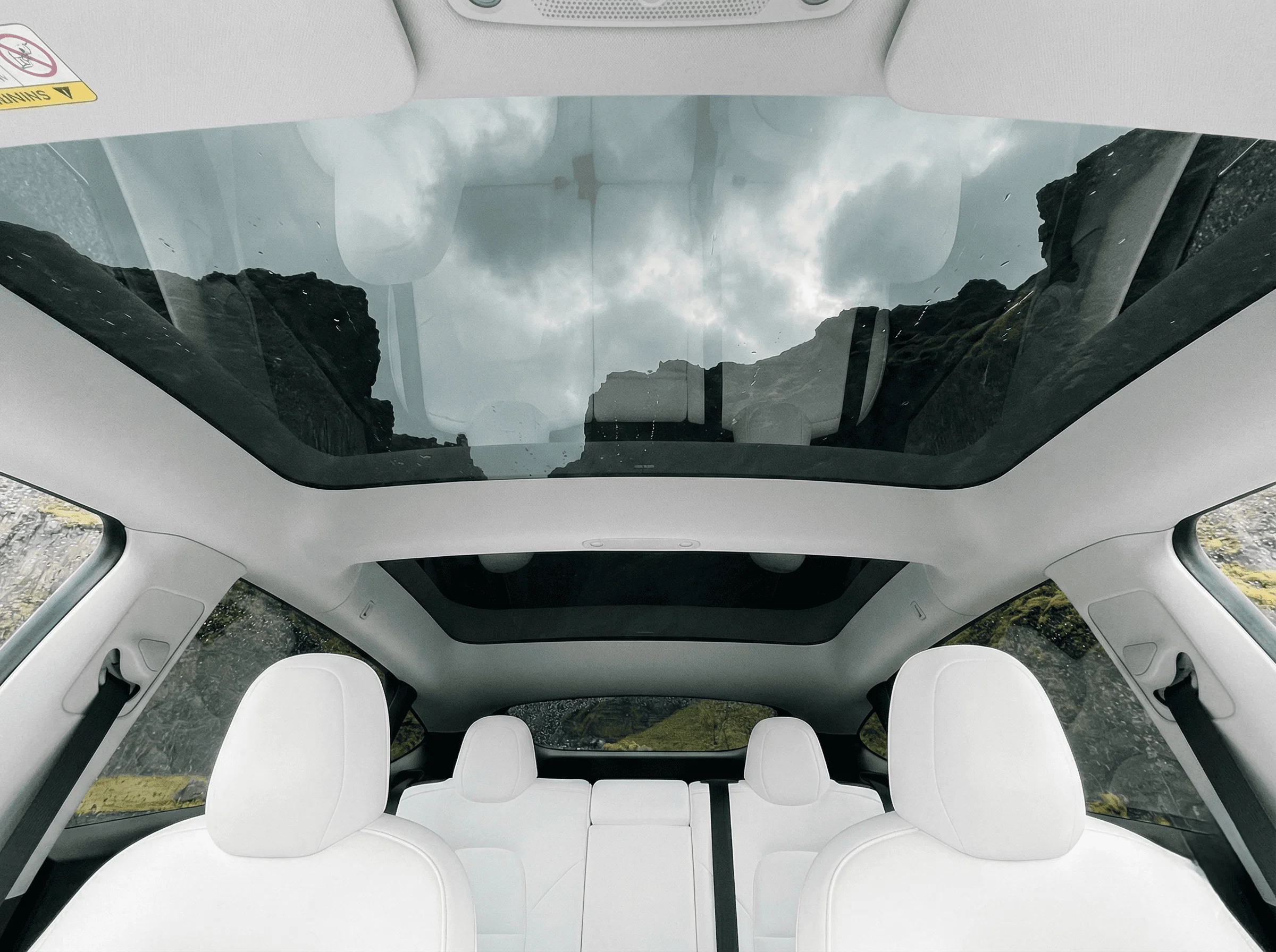 Low-angle interior view looking up through the panoramic glass roof of a car with white seats at a cloudy sky.