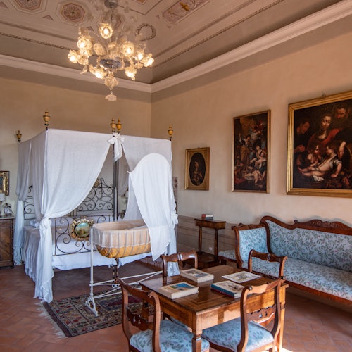 一间古色古香的房间，配有华盖床、婴儿摇篮、木制家具和墙上的经典画作，由精美的枝形吊灯照明。