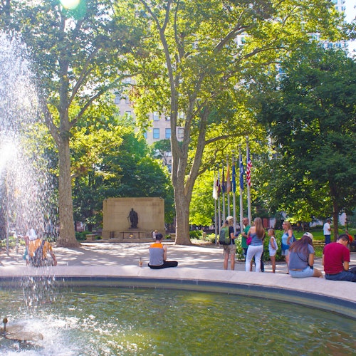 人们在一个阳光明媚的公园里放松，公园里有喷泉、旗帜、树木，背景则是一个雕像。