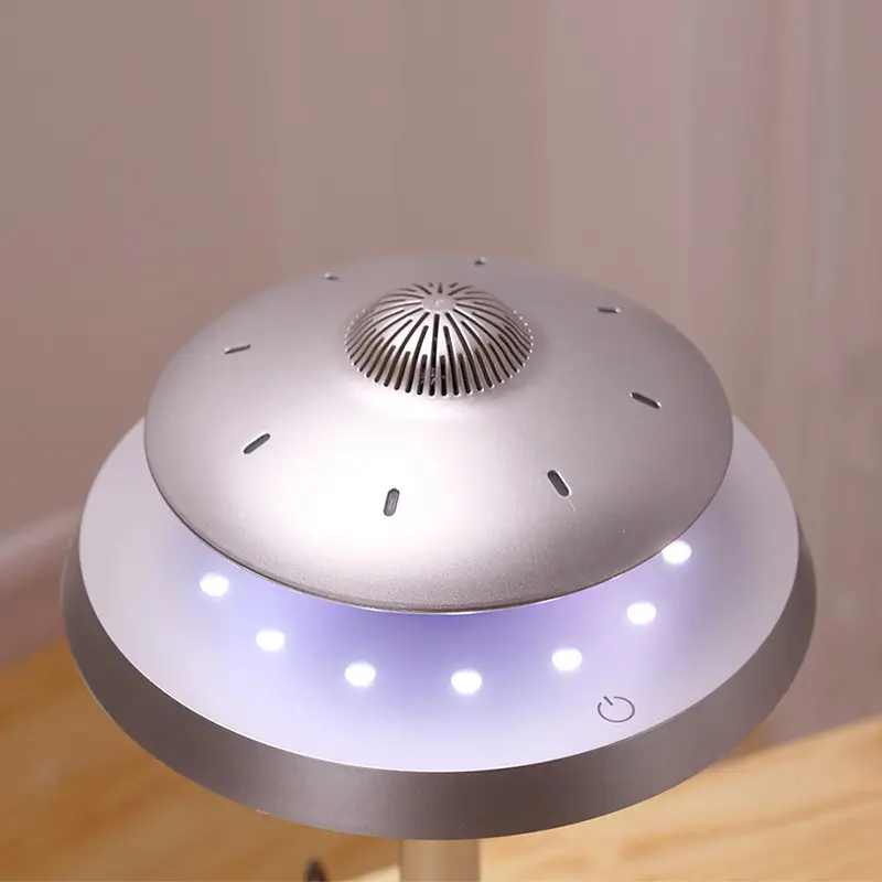 A modern, levitating, silver UFO-shaped lamp with a circular design and a central grille, surrounded by small LED lights, is placed on a wooden surface. The lamp features a power button on the lower edge.