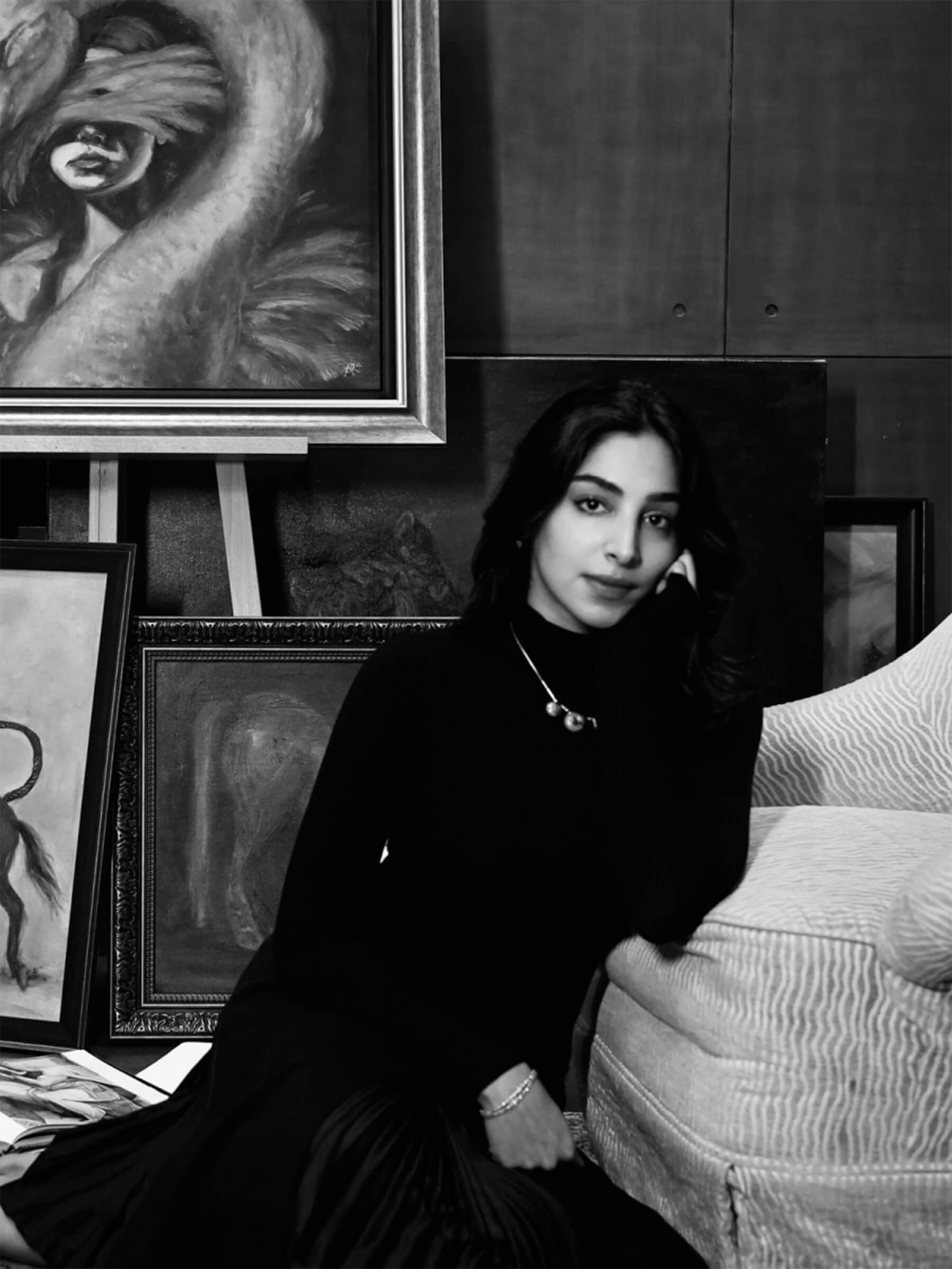 Black-and-white photo of a woman seated beside framed paintings in an art studio.