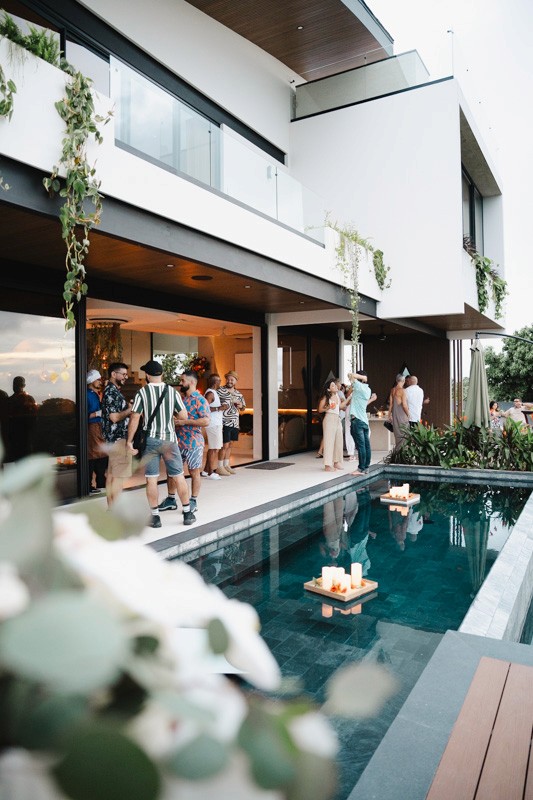 Group pool party at sunset on the deck of a modern multi-story luxury villa with floating candles in the pool.
