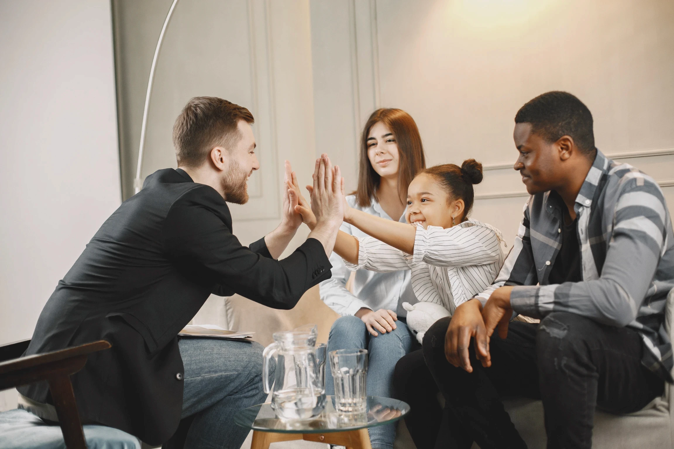 A group of four diverse friends sit smiling on a couch, high-fiving each other with enthusiasm. The room has a cozy, relaxed atmosphere.