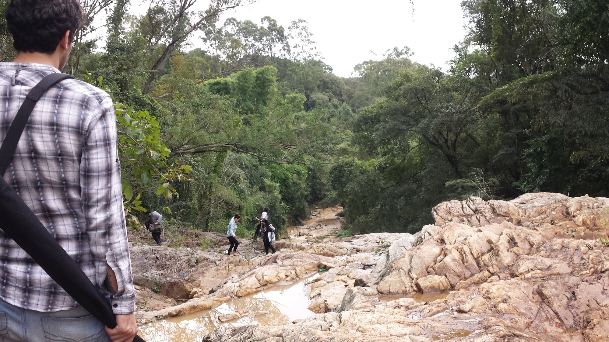 Equipe segue pelas pedras de um rio cercado de mata, com homem de mochila em primeiro plano