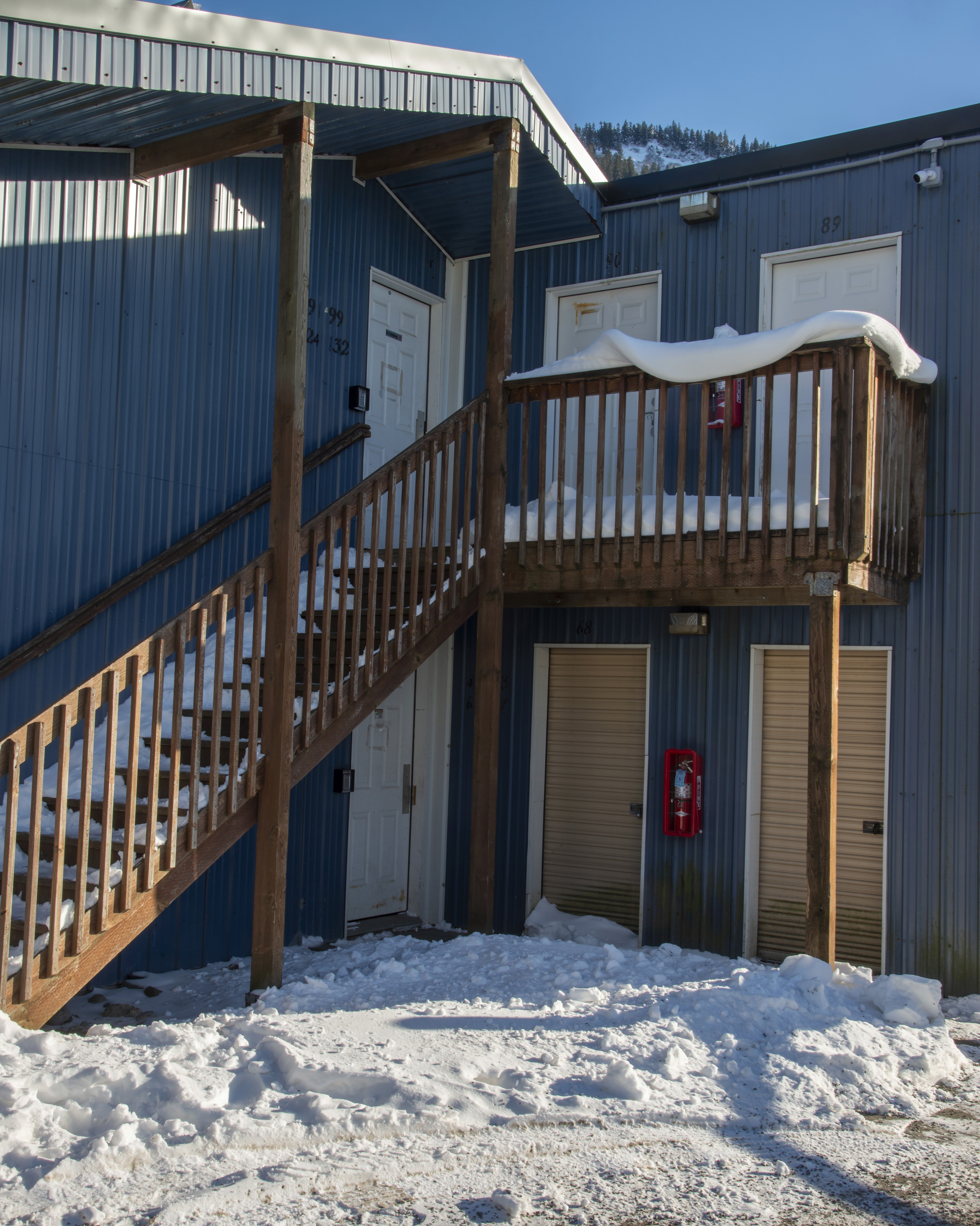 Snow-covered exterior staircase with wood railings leading to a second-floor landing on a blue metal panel storage building, with snow piled at the base and a clear winter sky in the background.