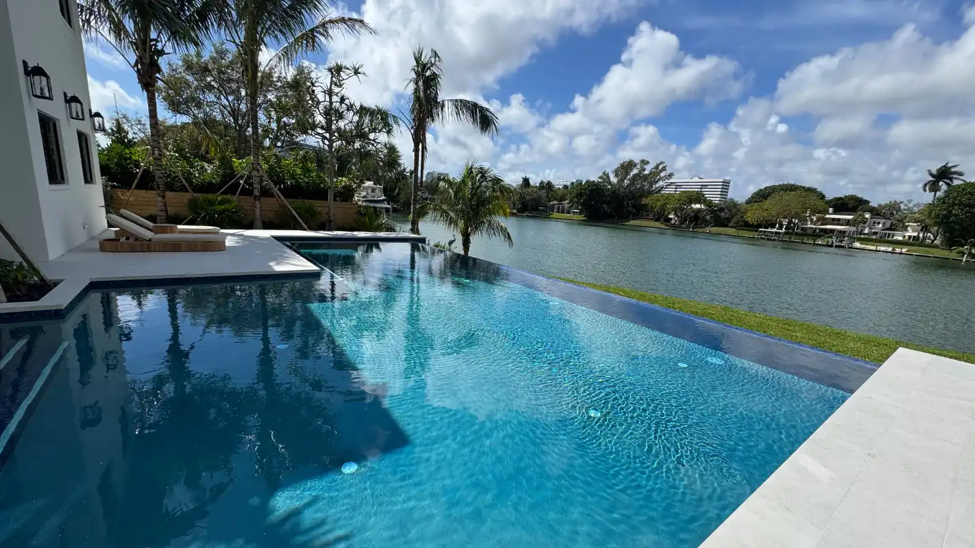  Infinity-edge pool at a Bay Point waterfront estate, overlooking a canal lined with palms and homes, with sun loungers on the deck and the Miami skyline in the distance.