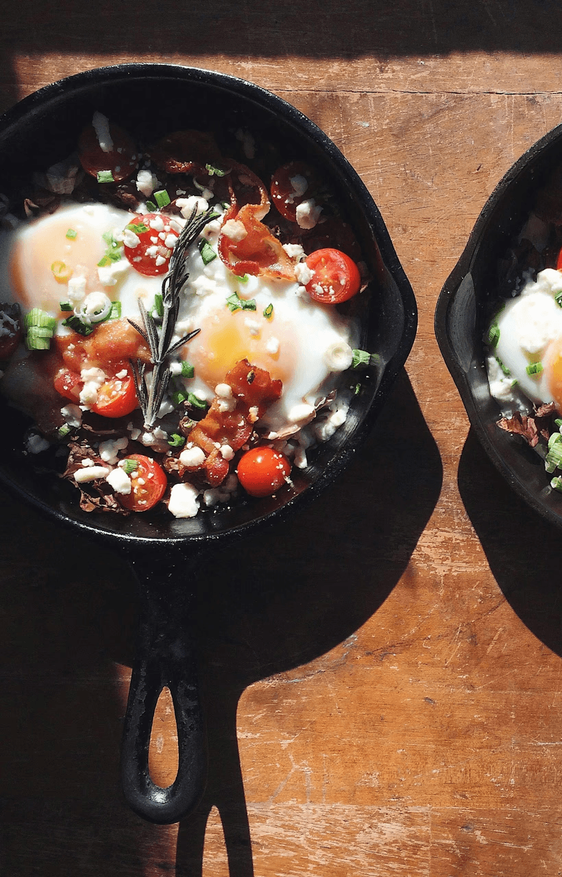 Deux œufs au plat dans une poêle en fonte, garnis de tomates cerises, feta, bacon, ciboulette et romarin, sur une table en bois.