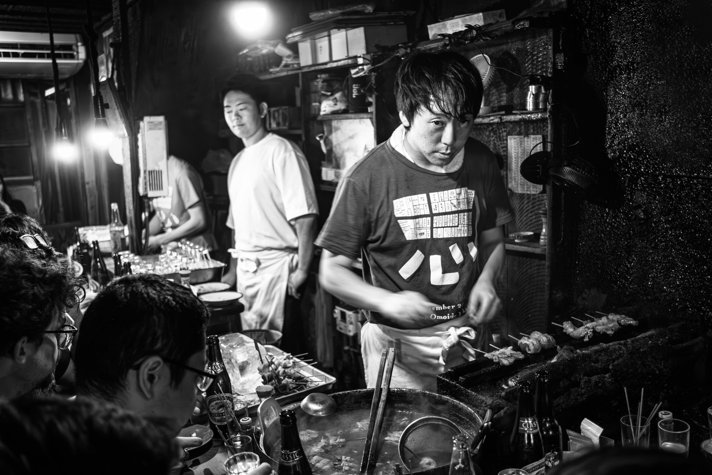 Black and white photograph of a crowded izakaya kitchen with a chef grilling skewers.