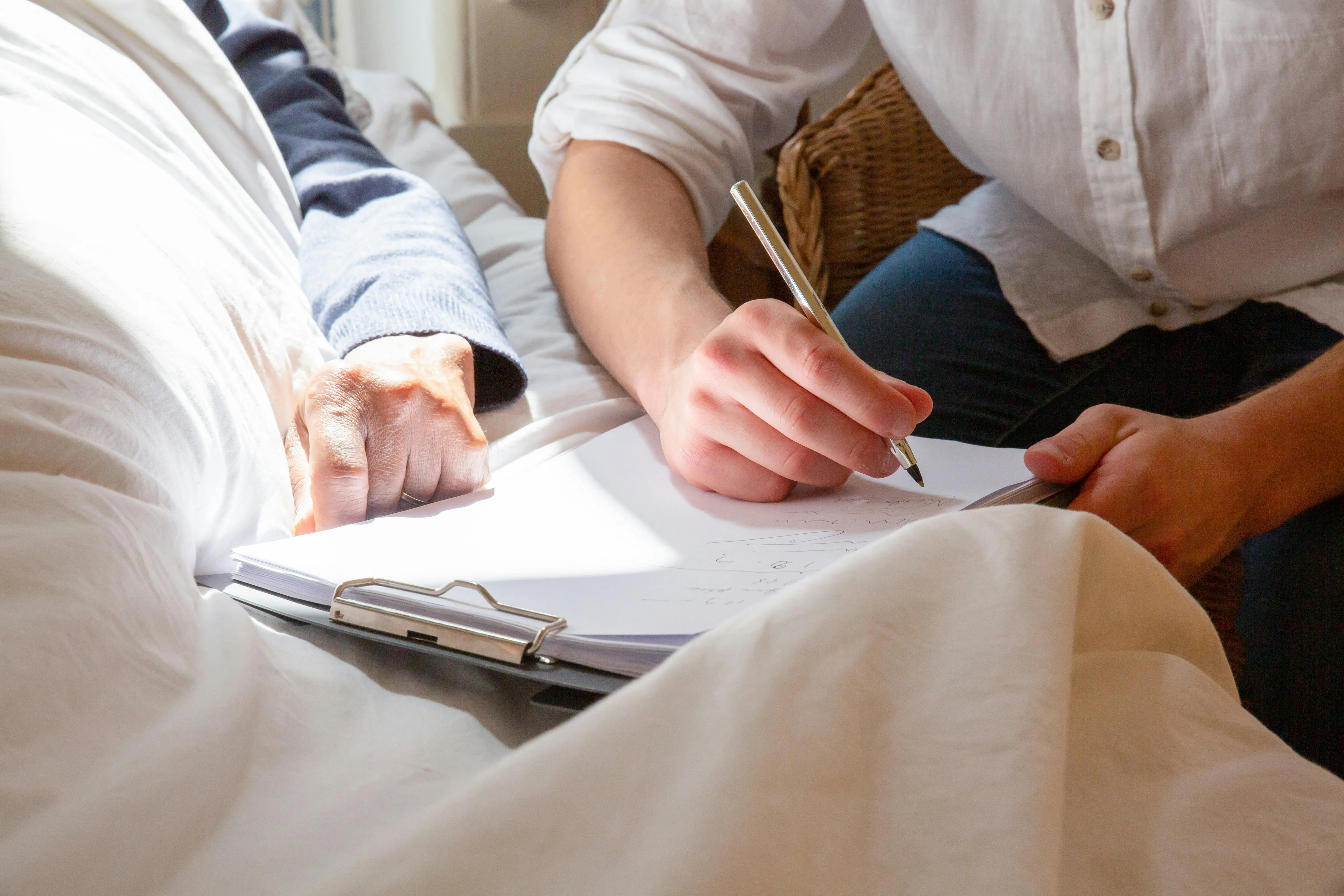 a death doula next to a sick person taking notes on a clipboard to create legacy projects, there's a lot of light and white colors