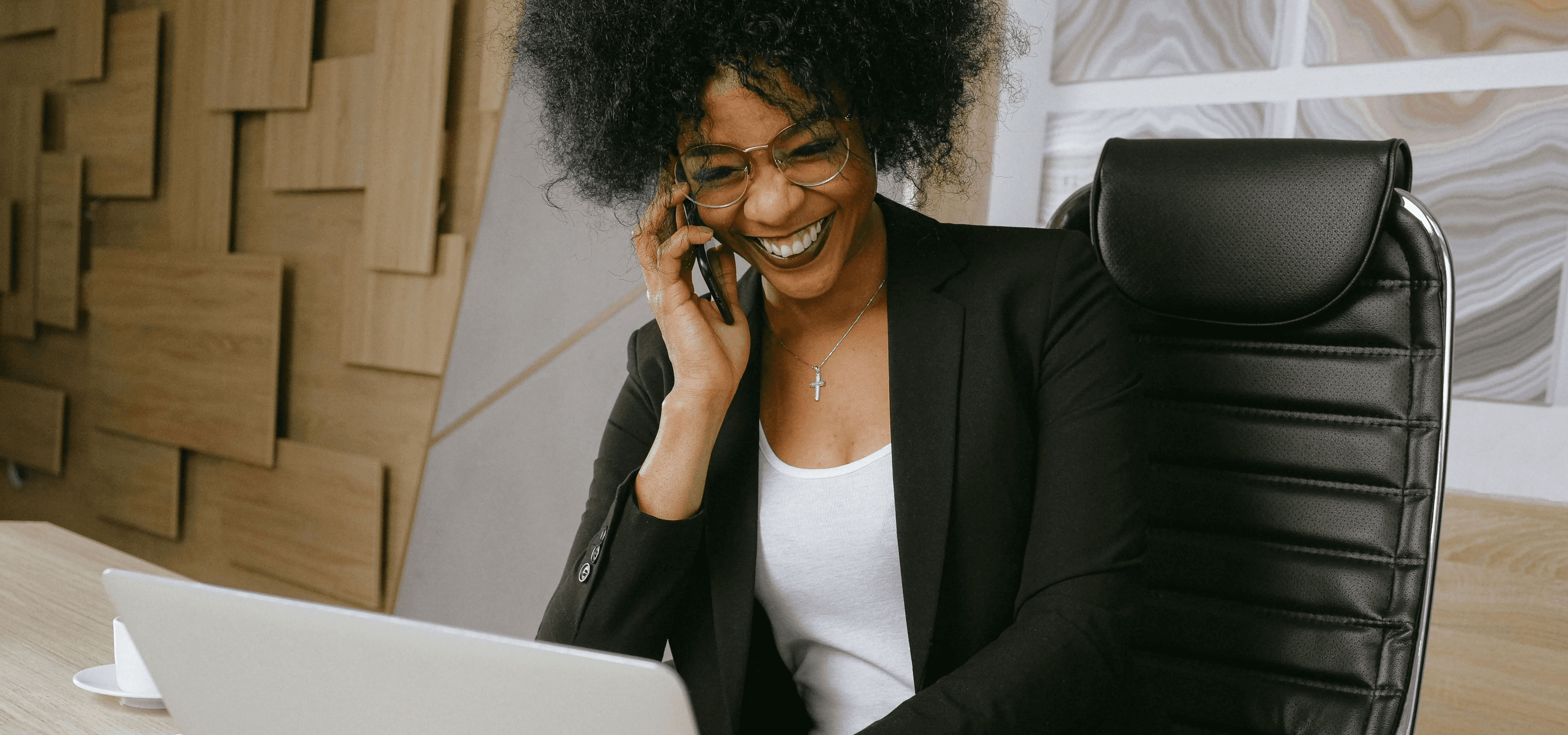 Professional woman smiling while talking on phone and working on laptop in modern office