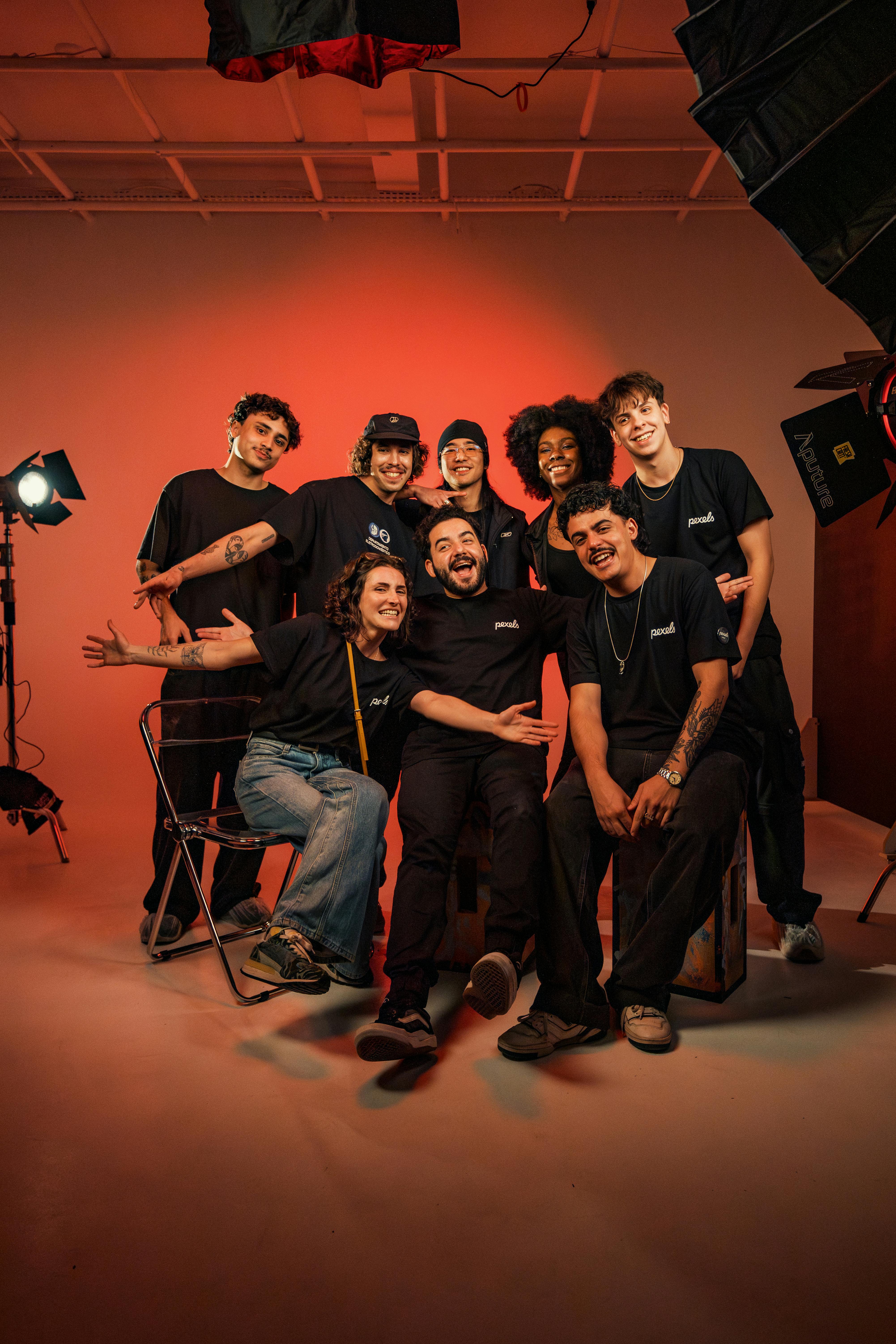Eight smiling young adults in black Pexels shirts pose in a red-lit studio with professional photography lighting gear.