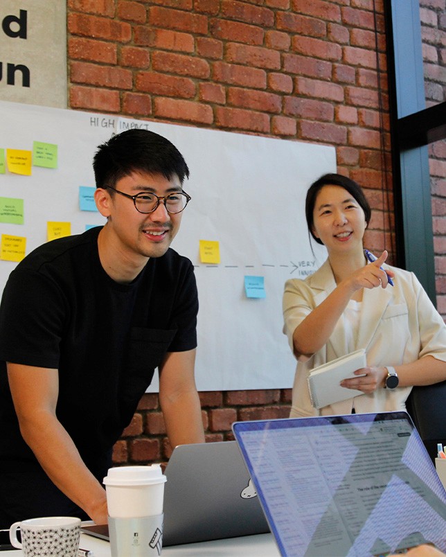 Two members of 55 Minutes collaborating in a modern workspace, discussing ideas in front of a laptop and a board with sticky notes.